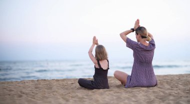 Mutlu aile annesi ve kızı yoga yapar, sahilde lotus pozisyonunda meditasyon yaparlar. Rahatlama, rahatlama ve doğayla uyum.