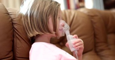 Evdeki kanepede nebulizör kullanan tıbbi oksijen maskeli hasta küçük kız. Çocuk tedavisinde nebulizatör solunumu