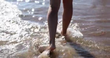 İnce kadın bacakları ve ayakları kumlu sahilde deniz suyunun dalgalarında yürüyor. Deniz sörfünde yürüyen güzel bir kadın