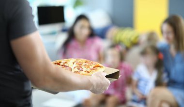 Erkek eller karton tezgahta pizza taşır. Arka planda, iki kadın ve iki çocuk kanepede oturuyor..