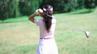 Takım elbiseli kadın arkasında golf sopası tutuyor. Yeşil çimlerde golf oynayan birinin arka planı.