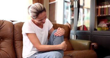 Oturma odasında kanepede oturan kadın dizlerinde ve bacaklarında ağrıdan muzdarip masaj, kas gerginliği ve tendon kasılması.