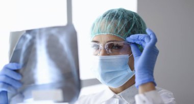 Doktor röntgen filmini inceliyor. Koruyucu maskeli ve takım elbiseli bir kadın gözlüğünü eliyle ıslatır ve ciğerlerinin fotoğrafını çeker..