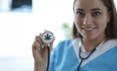 Close-up of female hand holding stethoscope with cannabis sign. Smiling doctor practitioner in medical gown. Medicine and healthcare. Medical marijuana concept clipart