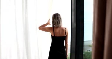 Siyah gömlekli güzel bir kadının arka manzarası yeni bir güne başlıyor ve perdeleri açıyor. Kız modern apartman veya otel odasındaki manzaraya panoramik pencereden bakar ve yatak odasında rahatlar.