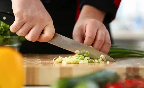 Cook holds knife in hand and cuts on cutting board green onions for salad or fresh vegetable soup with vitamins. Raw food and vegetarian recipe book in modern society popular concept.