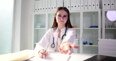 Genç doktor masada oturur elini ileri uzatır, yakın plan. Klinikte dostça karşılama.