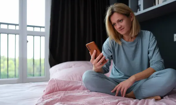 Young woman in pajamas sitting in bed with mobile phone in hand. Phone addiction nomophobia concept