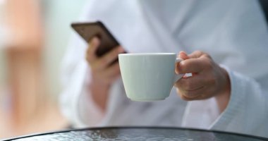 Cep telefonu ve bardak tutan terli bornozlu bir kadının kesilmiş fotoğrafı. Haberleri izlemek ve sabah kahvesini rahatlatmak.
