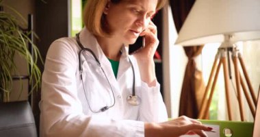 Woman doctor typing on laptop keyboard and talking on mobile phone 4k movie. Remote medical consultation concept