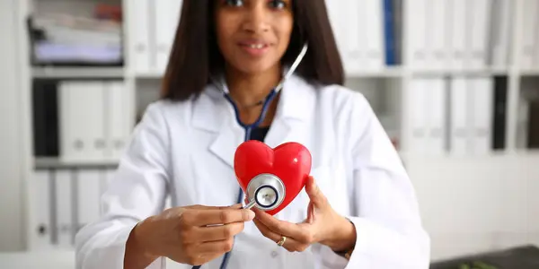 stock image Beautiful black smiling female doctor hold in arms red toy heart closeup. Cardio therapeutist student education CPR 911 life save physician make cardiac physical pulse rate measure arrhythmia