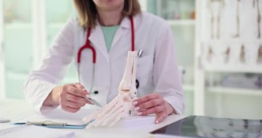Kadın doktor, hafif hastane önermesi üzerine masada oturan yapay ayak iskeleti modelinde kemikleri kalemle işaretliyor. Kadın insan eklemlerinin yapısını yavaş çekimde gösterir