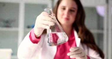 Lastik eldivenli genç bir kadın cam şişeyi temiz su numunesiyle tutuyor. Bilim adamı, laboratuvarda yavaş çekimde şeffaf sıvının durumunu inceliyor.