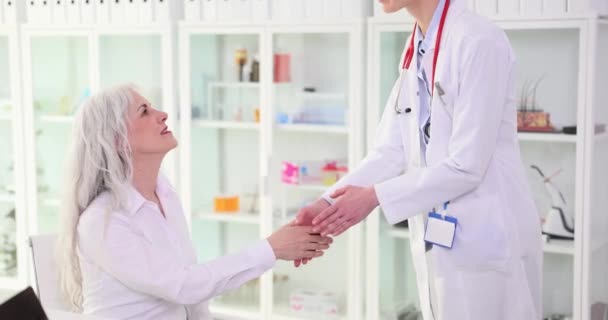 ブロンドの医者はクリニックのオフィスで成熟した女性クライアントの手を振る 感情的な女性の笑顔を迎える フレンドリーな挨拶と患者治療のコンセプトスローモーション — ストック動画