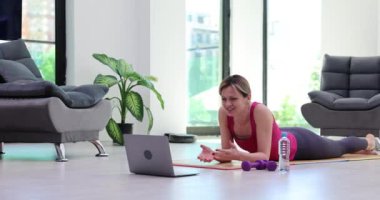 Spor giyinen formda bir kadın yoga minderine uzanıyor ve spor eğitmeniyle konuşuyor. Çevrimiçi ve uzak spor eğitimi video konferansı öğrenimi