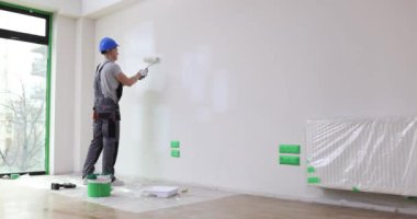 Miğferli ve tulumlu bir inşaatçı evde tekerlekli sandalyeyle duvarı boyuyor. İşi bitirme ve resim hizmetleri