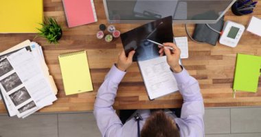 Hastanın eklem röntgenini inceleyen odaklanmış doktor tıbbi tarih ve üst görüş üzerine notlar alıyor. Klinikteki masada insan kemiğinin röntgenine bakan erkek ortopedi uzmanı.