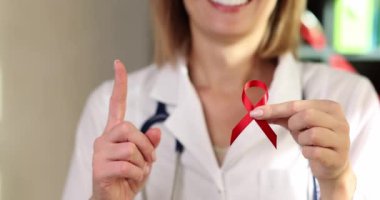 Beyaz laboratuvar önlüklü kadın doktor AIDS 'le mücadelenin kırmızı kurdelesini tutuyor ve parmağını kaldırıp gülümsüyor. HIV farkındalığı ve hastalıkların önlenmesi kavramı