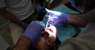 Dişçi, hastanın ağzında şırıngayla anestezi iğnesi yapar. Işıkla aydınlatır. Doktor, dişçi kliniğinde kadını ameliyata hazırlıyor.