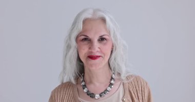 Stüdyoda kameraya poz veren gevşek gri saçlı ve kolyeli mutlu, şık yaşlı kadın portresi. Çekici yaşlı bayan beyaz ağır çekimde gülümser.