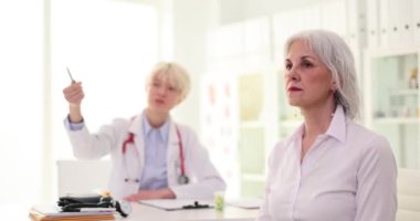 Oftalmolog, olgun bir kadının görüşünü kontrol etmek için Snellen dosyasına kalemle işaret ediyor. Kadın hasta avuçlarını yavaş çekimle kapatıyor.