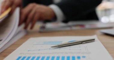Kalem, ofis masasında belge arayan müdüre karşı açılan soruşturma kağıdının üzerinde. Yavaş çekimde istatistik ve grafiklerle muhasebe malzemeleri