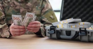 Askeri memur, para yığınlarıyla dolu açık bir davanın yanında duran dolarları sayıyor. Üniformalı adam ağır çekimde silah almak için parayı kontrol ediyor.