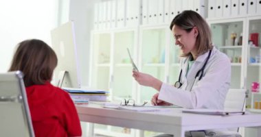 Gülümseyen kadın çocuk doktoru, iş yerindeki küçük hastanın test sonuçlarına bakıyor. Kadın ve kız klinikteki masada ağır çekimde oturuyor.