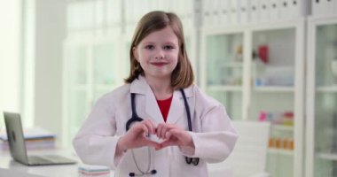 Beyaz önlüklü liseli kız doktor rolü yapıyor ve kalp işareti gösteriyor. Steteskoplu pozitif kız modern ofiste ağır çekimde duruyor.