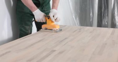 Yetenekli marangoz ahşap masa yüzeyini öğütücü makinesiyle parlatır. Profesyonel üniformalı bir erkek ahşap işlemek için ekipman kullanır.