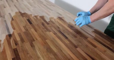 Erkek marangoz ahşap ofis masasının yüzeyini özel cila ile kaplar. İş üniformalı bir adam sıvıyı ağır çekimde bez parçasıyla süpürüyor.