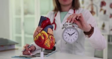 Kadın doktor ışık hastanesi ofisinde bilimsel kalp modelinin yanında çalar saati tutuyor. İnsan organının detaylı anatomisi ve yavaş çekim konsepti