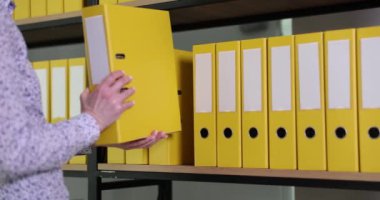 Kadın çalışan, muhasebe belgeleriyle birlikte sarı yüzük klasörünü ofis arşivine geri koyuyor. Rafta malzemeler olan organize dizin yapısı