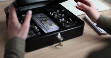Kadın, muhasebeci kasasını güvenlik kilidi ve dolar banknotlarıyla dolu bir anahtarla açıyor. Üzerinde banknotlar ve madeni paralar olan kara kutu.