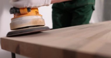 Marangoz yeni ahşap yüzeyin kenarlarını öğütme makinesiyle parlatır. Lastik eldivenli işçi, müşteriler için özenle yeni bir masa hazırlıyor.