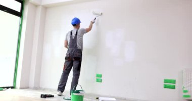 Foreman silindir kullanarak duvarı beyaza boyuyor. Miğferli ve profesyonel üniformalı işçiler, ev sahiplerinin daire arka görünümünde tadilat yapmasına yardım ediyor.