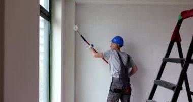 Miğferli ve tulumlu genç bir erkek işçi, duvarları tekerlekli fırçayla boyuyor. Profesyonel müteahhit, odanın iç tarafını yavaş çekimde yeniliyor
