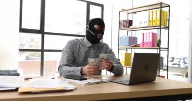 Hırsız maskeli heyecanlı adam ofiste bilgisayarla masada oturan kulaklıklı mikrofona konuşur. Duygusal hacker sinirli omuz silkerek para sayar
