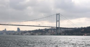 Şehirdeki geniş nehir boyunca hareket eden otomobilleri olan büyük, modern bir köprü. Martı yağmurdan önceki bulutlu yaz gününde dalgalı suyun yüzeyinde uçar.