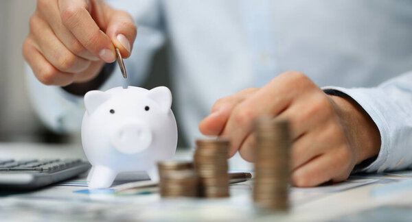 Man hand putting coin into white piggy bank closeup. Accumulation of money for investment in business concept