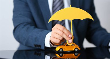 Takım elbiseli işçinin yakın çekimi sarı araba modelinin üzerinde ahşap şemsiye taşıyor. Araba korumasının sembolü. Otomobil sigortası, güvenlik, ehliyet kavramı