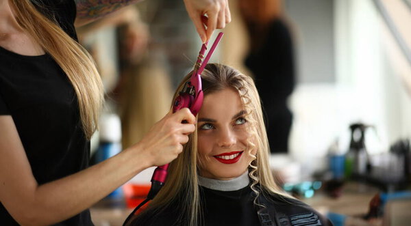 Portrait of blonde female on hairdresser appointment, master create curly hairstyle. Barber use curling iron tool for waving hair. Beauty salon concept