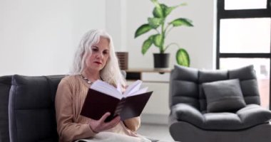 Konsantre olmuş olgun bir kadın rahat koltukta oturan kalın kitapların sayfalarını çevirir. Gri saçlı bayan hafta sonları evde kitap okumayı sever.