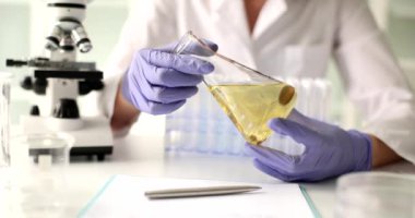 Bilim adamı elinde plastik eldivenlerle elinde ayçiçeği yağı bulunan cam matarayı tutuyor. Kadın laboratuvarda ağır çekimde sarı sıvı saflığı inceliyor.