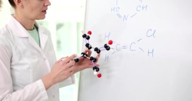 Bilim adamı, beyaz tahtaya yazılan kimyasal formülü eldeki molekül modeliyle karşılaştırarak açıklıyor. Laboratuvar önlüklü kimyager yavaş çekim semineri veriyor.