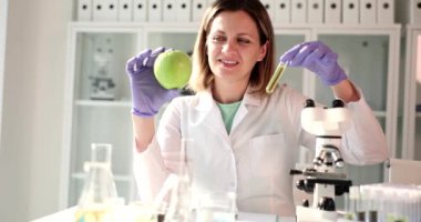 Pozitif bilim adamı, ellerinde taze elma ve sarı sıvıyla test tüpüne bakar. Kadın mikroskobun yanında ağır çekimde oturuyor.