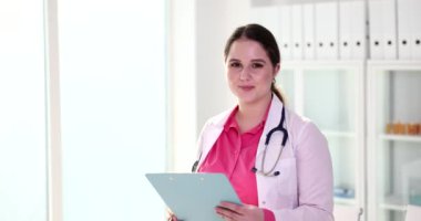 Steteskoplu genç doktor, panoda tedavi planını okuyor. Pozitif kadın kameraya bakar ve klinikte ağır çekimde gülümser.