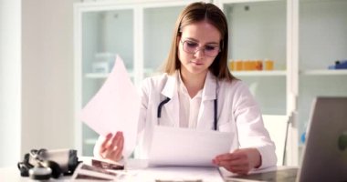 Odaklanmış doktor, dizüstü bilgisayarına bakan hasta materyallerini inceliyor. Gözlüklü genç kadın tıp tarihi okuyor. Masada ağır çekimde oturuyor.