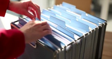 Sekreter analiz için muhasebe belgelerini içeren gerekli klasörü arıyor. Kadın ofis çekmecesindeki organize arşiv malzemelerini yavaş çekimde kontrol ediyor.