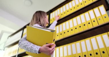 Sekreter evrak işlerini halletmek için üst rafa uzanıyor. Ofiste ağır çekimde arşiv materyalleri olan düzenli sarı klasörler